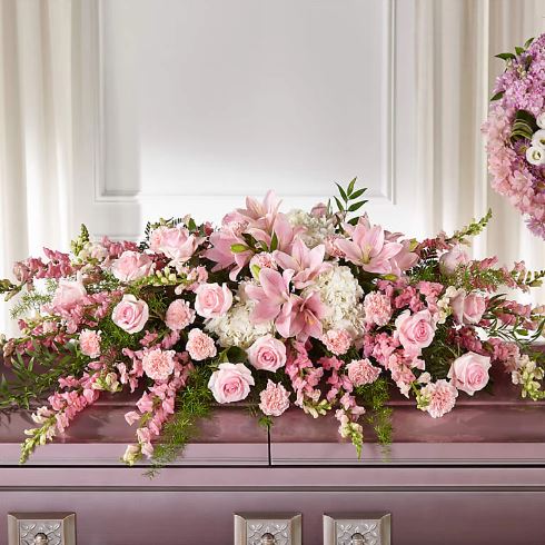 A premium casket spray floral arrangement featuring soft pink lilies, roses, carnations, and white hydrangeas, elegantly draped over a lavender-colored casket.