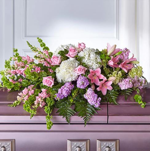 A large, elegant casket spray floral arrangement featuring a mix of white hydrangeas, pink roses, pink lilies, and purple stock flowers, accented with green Bells of Ireland and lush ferns, resting on a polished lavender-colored casket.