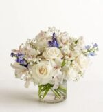 A lush floral arrangement in a low glass cylinder vase featuring white roses, white alstroemeria, baby's breath, and accents of purple delphinium.