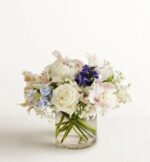 A compact floral arrangement in a clear cylindrical glass vase featuring a large white rose, white alstroemeria with pink accents, light blue delphinium, purple stock flowers, and white baby's breath against a plain white background.