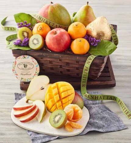 A premium dark wood woven basket filled with assorted fresh fruits including a large mango, pears, apples, kiwis, and oranges, decorated with purple flowers and a green brand ribbon, accompanied by a white plate of sliced fruit in the foreground.
