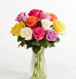 A premium floral arrangement featuring one dozen assorted roses in vibrant shades of pink, yellow, orange, white, and lavender, presented in a clear fluted glass vase against a minimalist white background.