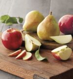 A red apple and two green pears on a rustic wooden cutting board, with various fruit slices and green leaves scattered around.