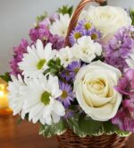 A beautiful floral arrangement in a wicker basket featuring white roses, white daisies, and purple accent flowers like alstroemeria and asters.
