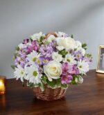 A lush floral arrangement in a woven brown wicker basket featuring white roses, white daisies with yellow centers, lavender stock, and purple alstroemeria, set on a dark wooden table.