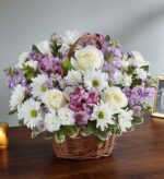 A premium floral arrangement in a woven brown wicker basket featuring white roses, white daisies, lavender alstroemeria, and purple stock flowers, set against a neutral background.
