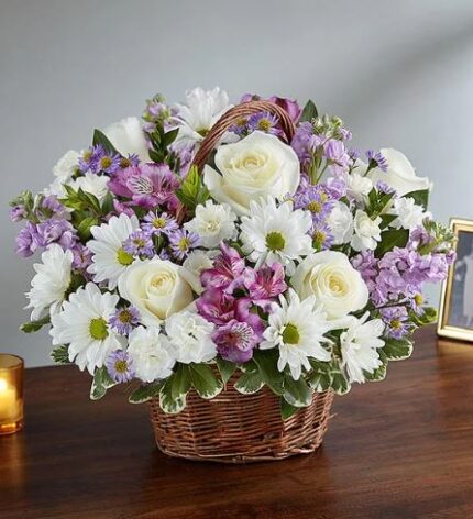 A premium floral arrangement in a woven brown wicker basket featuring white roses, white daisies, lavender alstroemeria, and purple stock flowers, set against a neutral background.
