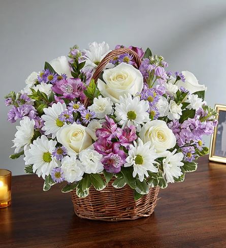 A premium floral arrangement in a woven brown wicker basket featuring white roses, white daisies, lavender alstroemeria, and purple stock flowers, set against a neutral background.