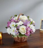A "Peace, Prayers & Blessings" floral arrangement featuring white roses, white daisies, and purple alstroemeria in a small woven brown basket on a wooden table.