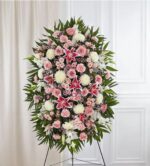 A large, elegant oval-shaped funeral standing spray featuring a dense arrangement of pink roses, stargazer lilies, white chrysanthemums, and pink carnations accented with lush green palm fronds and filler flowers on a wire easel.