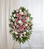 A large, vertical funeral standing spray arrangement featuring pink lilies, pink roses, white chrysanthemums, and pink carnations accented with lush green palm leaves on a wire easel.
