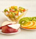 A clear glass bowl filled with fruit salad containing chopped kiwi, oranges, and apples, accompanied by plates of sliced red pears, kiwi rounds, and orange slices on a white wooden surface.