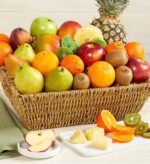 A large, overflowing woven wicker basket filled with a variety of fresh fruits including apples, pears, oranges, mangoes, kiwis, and a whole pineapple, with sliced fruit samples on the side.