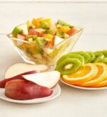 A glass bowl filled with a colorful fruit salad alongside plates of sliced red pears, green kiwis, and orange rounds on a light wooden surface.