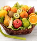 A woven wicker gift basket overflowing with a variety of fresh orchard fruits, including apples, pears, oranges, and sliced kiwis, nestled on green tissue paper with a lime green ribbon.
