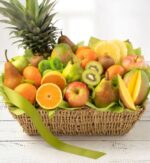 A lush, overflowingly abundant woven fruit basket featuring a whole pineapple, various pears, mangoes, apples, kiwi slices, and halved oranges, decorated with a long lime-green ribbon on a white wooden surface.