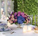A lush floral centerpiece featuring purple roses, blue hydrangeas, and light pink spray roses in a silver pedestal bowl on a set wedding table.