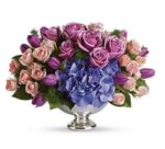 A lush floral arrangement featuring purple roses, pink spray roses, blue hydrangea, and purple tulips in a silver pedestal bowl.