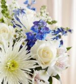A lush floral arrangement featuring white roses, white chrysanthemums, and vibrant blue delphiniums accented with white alstroemeria and small aster flowers.