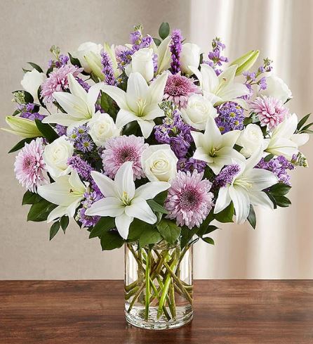 A large, premium floral arrangement in a clear glass cylinder vase, featuring a mix of white lilies, white roses, lavender chrysanthemums, and purple stock flowers against a neutral background.