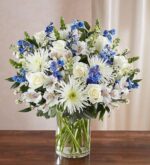A large, elegant floral arrangement in a clear glass cylinder vase featuring white roses, white spider mums, blue delphinium, and white alstroemeria with green foliage.