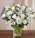 A serene floral arrangement in a glass cylinder vase featuring white roses, white spider mums, blue delphiniums, and white alstroemeria with green foliage.