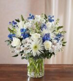 A lush floral arrangement in a clear glass cylinder vase featuring white roses, white spider mums, blue delphinium, and white alstroemeria with green foliage, set against a neutral backdrop on a wooden surface.
