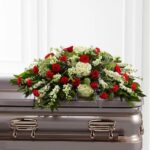 A deluxe Sincerity Casket Spray featuring a lush arrangement of red roses, red carnations, white hydrangeas, and white snapdragons with green foliage on a metallic casket.
