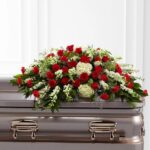 A premium funeral casket spray featuring a dense arrangement of vibrant red roses, red carnations, white hydrangeas, and white snapdragons nestled in lush green foliage, resting atop a polished metallic casket.