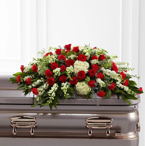 A premium funeral casket spray featuring a dense arrangement of vibrant red roses, red carnations, white hydrangeas, and white snapdragons nestled in lush green foliage, resting atop a polished metallic casket.