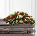 A large, elegant casket spray floral arrangement featuring vibrant red roses, white hydrangeas, and white snapdragons with lush green foliage, resting on a silver-toned metal casket.