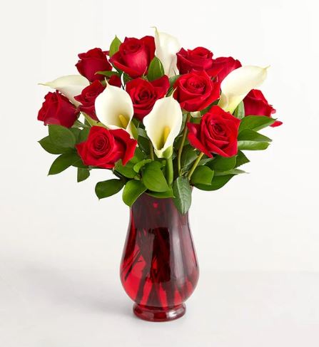 A vibrant bouquet of deep red roses and elegant white calla lilies arranged with lush green leaves in a translucent ruby-red glass vase.