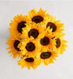 A top-down view of a vibrant "Deluxe" sunflower arrangement featuring approximately ten large, golden-yellow sunflowers with dark brown centers, tightly bunched together against a light gray marbled background.