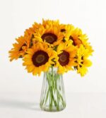 A lush and dense bouquet of bright yellow sunflowers with dark brown centers, elegantly arranged in a clear, ribbed glass vase against a minimalist white background.