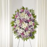 A large, elegant funeral standing spray arrangement featuring a blend of white lilies, lavender roses, and purple accent flowers, framed by lush green palm fronds on a wire easel.