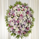 A large, oval-shaped funeral standing spray composed of pink roses, white lilies, white daisies, and purple stock flowers, accented with lush green palm fronds and emerald foliage. A large, oval-shaped funeral standing spray composed of pink roses, white lilies, white daisies, and purple stock flowers, accented with lush green palm fronds and emerald foliage.