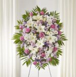 A large, elegant funeral standing spray titled "Surrounded by Love," featuring white lilies, pink roses, white daisies, and purple stock accented with lush green palm fronds on a wire easel.