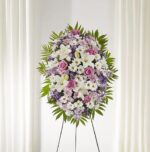 A vertical funeral standing spray floral arrangement on a wire easel, featuring a mix of white lilies, pink roses, white daisies, and purple accent flowers surrounded by green palm fronds.