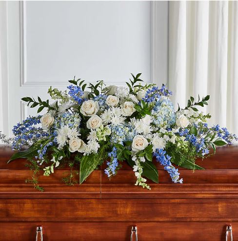 A premium "Timeless Essence" casket spray floral arrangement featuring white roses, blue hydrangeas, white lilies, and blue delphiniums, elegantly draped over a polished mahogany wooden casket.