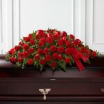 A deluxe "Tribute of Life" casket spray featuring a lush arrangement of vibrant red roses and carnations accented with green foliage and elegant red ribbons, resting on a dark wood casket.
