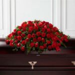 A lush and large "Tribute of Life" premium casket spray featuring a dense arrangement of vibrant red roses and carnations accented with green foliage and a red satin ribbon on a dark wood casket.
