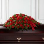 A large, elegant floral casket spray consisting of vibrant red roses and carnations accented with green foliage and a red ribbon, resting on a dark wood casket.