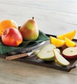 A rustic wooden tray featuring a whole red apple, a whole pear, and an orange in a dark bowl, accompanied by sliced pieces of the same fruits and a small wooden-handled knife on a light wooden surface.