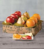 A Harry & David "Triple Treat" fruit gift box containing red apples, Royal Riviera pears (one wrapped in gold foil), and large oranges, arranged with green tissue paper in a decorative cardboard box.