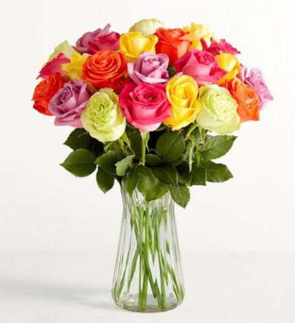 A premium floral arrangement featuring one dozen assorted roses in vibrant shades of pink, yellow, orange, white, and lavender, presented in a clear fluted glass vase against a minimalist white background.