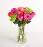 A vibrant bouquet of pink gerbera daisies, orange roses, and green chrysanthemums in a clear fluted glass vase.