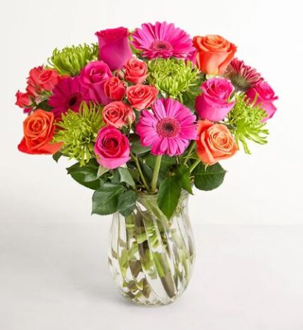 A vibrant floral arrangement featuring hot pink gerbera daisies, orange roses, pink roses, and lime green button poms in a clear glass spiral-textured vase.