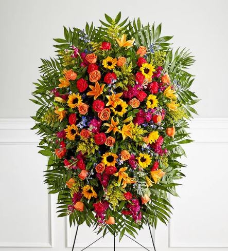 A large, extra-premium funeral standing spray featuring a vibrant mix of sunflowers, orange roses, lilies, and magenta carnations set against lush emerald palm fronds on a wire easel.