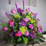 A vibrant and lush floral arrangement featuring purple roses, pink carnations, and green chrysanthemums accented with tall purple Liatris spikes and green ferns.