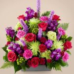 A lush and vibrant "Warm Embrace" premium floral arrangement featuring deep pink roses, purple stock flowers, lime green spider mums, and pink carnations accented with green hypericum berries and lush ferns in a sleek gray rectangular container.